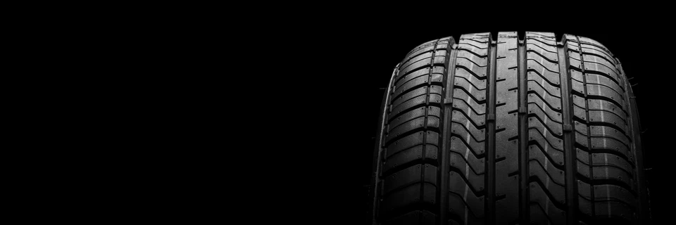 Gros plan artistique sur un pneu automobile neuf avec profil de bande de roulement détaillé, photographié sur fond noir avec éclairage dramatique latéral créant des contrastes marqués