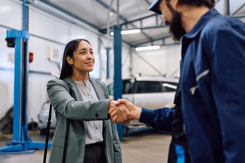 Mécanicien automobile en uniforme bleu avec badge nominal discute avec cliente blonde devant garage en briques rouges