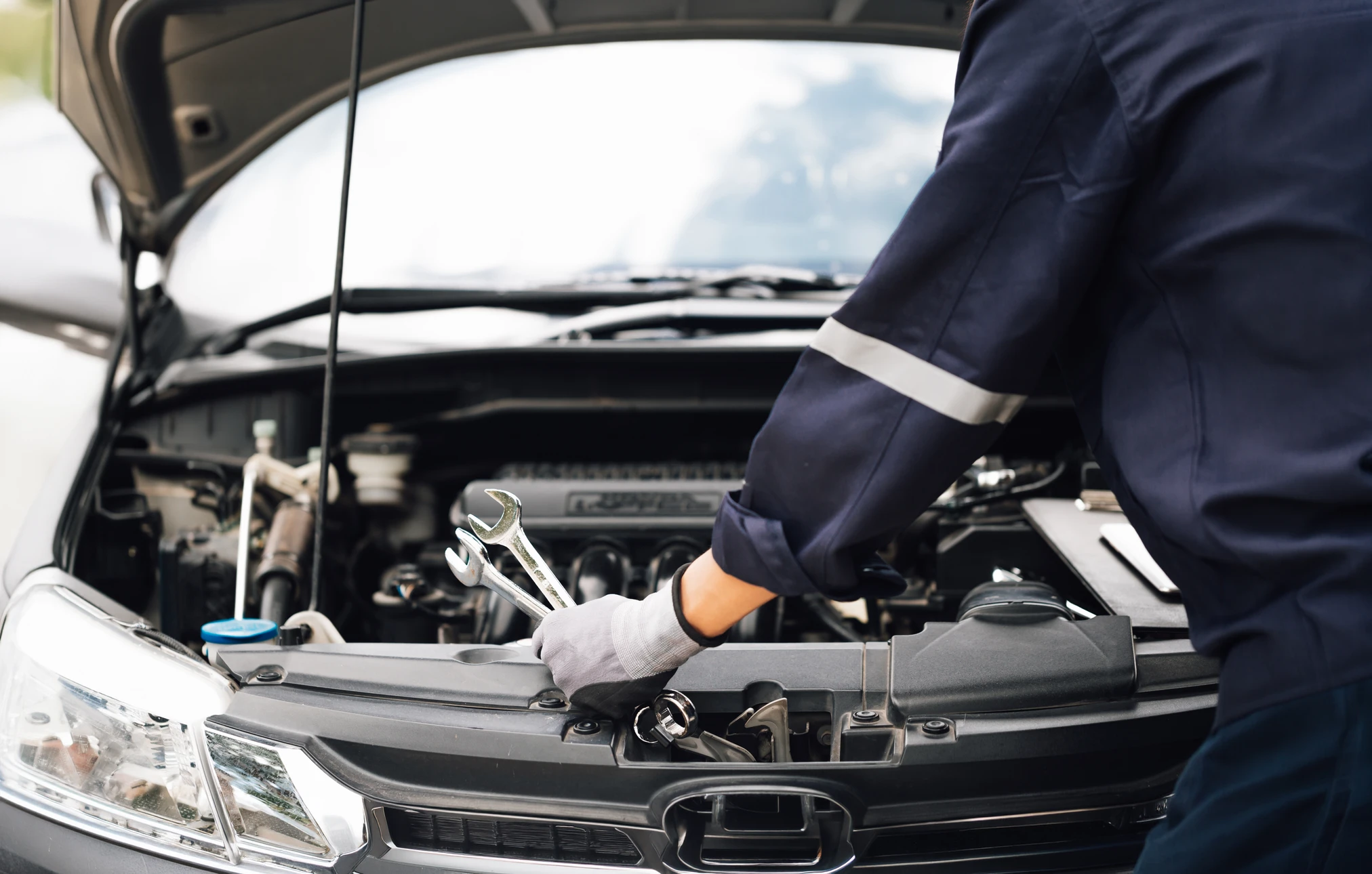 Atelier de réparation automobile avec mécanicien en combinaison bleue travaillant sous le capot d'une berline grise surélevée sur pont élévateur hydraulique